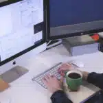 Person working on a dual monitor setup with email communications displayed, holding a green coffee mug.